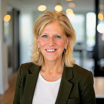 A professional headshot of a smiling woman with shoulder-length blonde hair. She is wearing a dark olive green blazer over a white top, a thin gold necklace, and gold hoop earrings. The background is a softly blurred modern office setting with warm, circular bokeh lights.