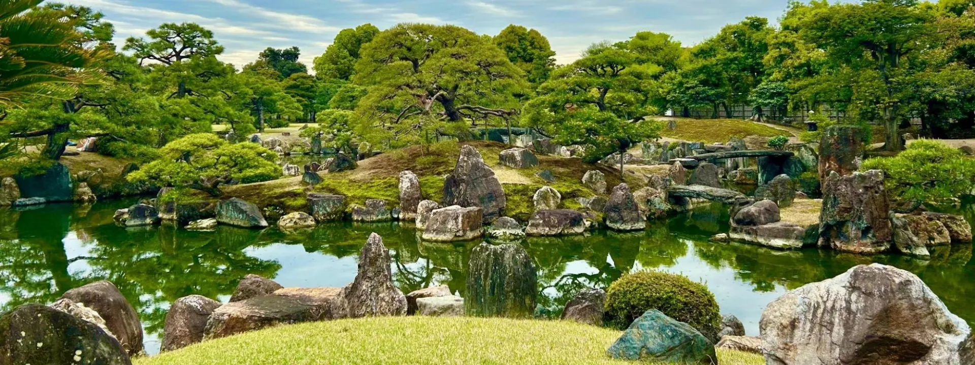 A traditional East Asian garden featuring a tranquil green pond surrounded by mossy rocks, carefully pruned pine trees, grassy mounds, and a stone bridge under a partly cloudy sky.