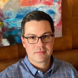 Portrait of a man with short dark hair, black-rimmed glasses, and a light blue and pink patterned button-up shirt, seated in front of a colorful abstract painting.