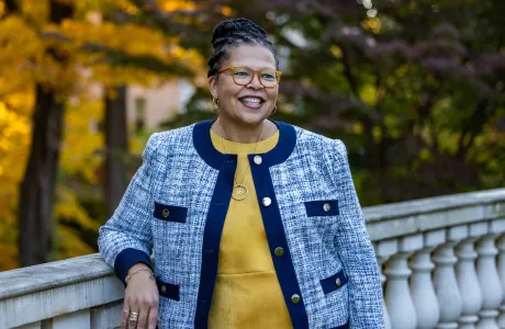 Sarah Willie-LeBreton smiles and looks away from the camera, leaning on a railing outdoors, in front of fall foliage, wearing a yellow shirt with a blue jacket on top.