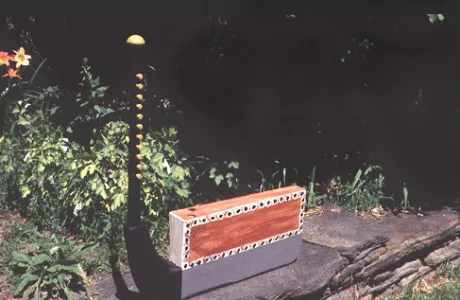 A harp with a patterned rectangular base sits on stone amidst lush greenery and flowers