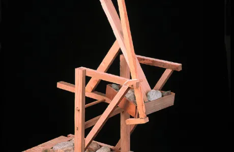 A wooden abstract sculpture is set against featuring angular beams and supports. Several rocks are placed within the structure.