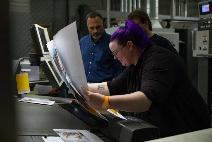 Three people standing around a table, looking closely at the printed sheets and smiling