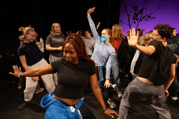 Several students dancing with their arms up in the air, a purple background behind them on a stage