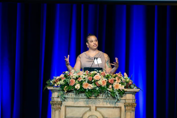 A speaker stands at a podium in front of a blue curtain, gesturing emphatically with her hands.