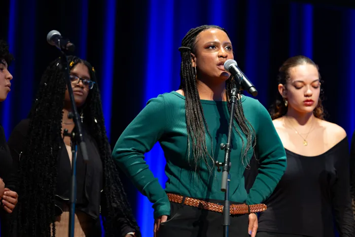 A student in a long-sleeved green shirt stands with her hands on her hips, singing into a microphone