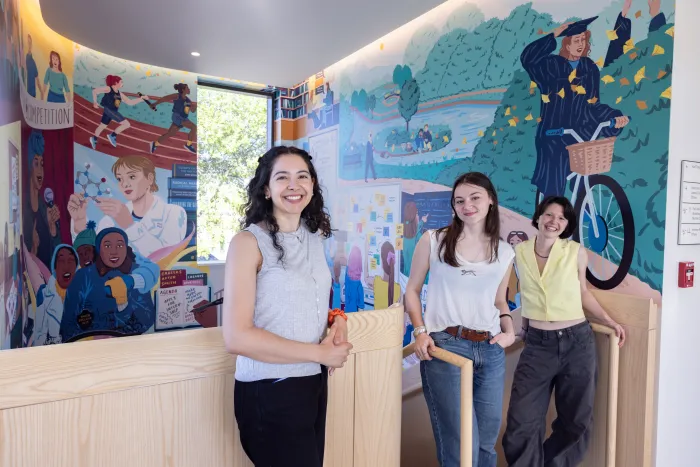 The two students and artist who worked on the mural in Kathleen McCartney Hall stand at the top of the stairs, smiling at the camera, with the mural behind them.