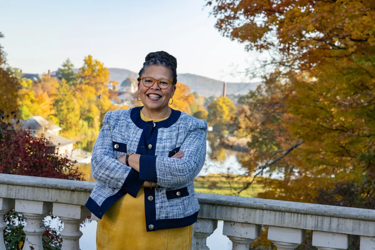 Sarah Willie-LeBreton smiles toward the camera in front of fall foliage, leaning on a railing and crossing her arms, wearing a yellow shirt with a blue jacket on top.