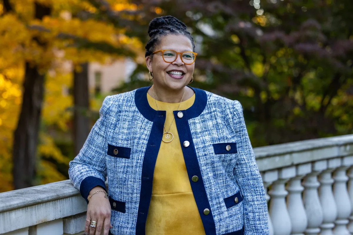 Sarah Willie-LeBreton smiles and looks away from the camera, leaning on a railing outdoors, in front of fall foliage, wearing a yellow shirt with a blue jacket on top.