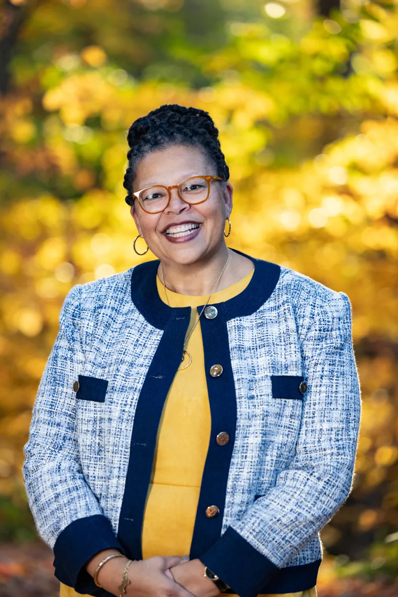 Sarah Willie-LeBreton smiles toward the camera in front of fall foliage, wearing a yellow shirt with a blue jacket on top.