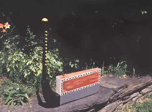 A harp with a patterned rectangular base sits on stone amidst lush greenery and flowers