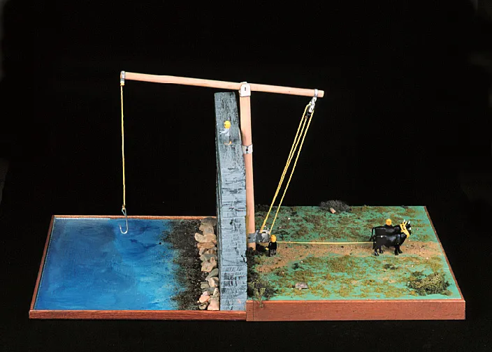A wooden lever balances between a water-filled area on the left and a green field with an ox on the right