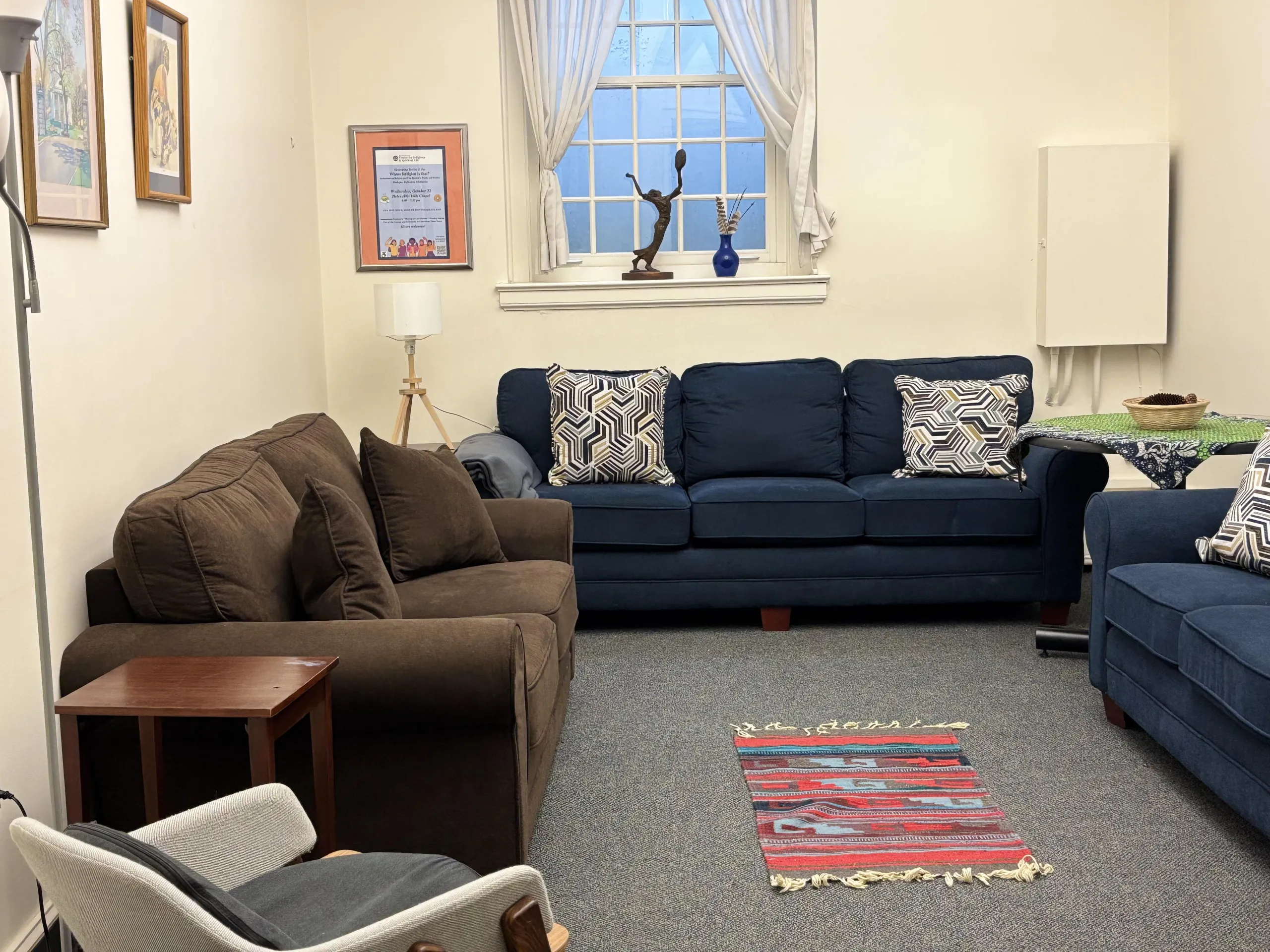 Cozy room with a brown sofa, two blue couches, patterned pillows, and a colorful rug. A window with curtains and framed art adds warmth.
