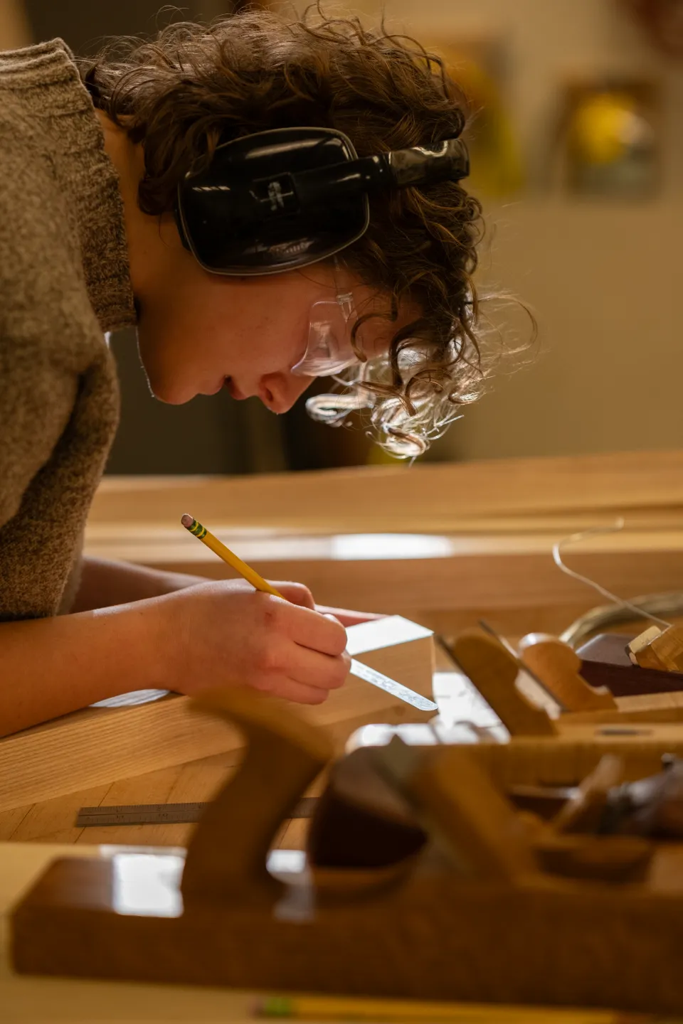 Eliza Berle ’27, wearing headphones, works on a piece of wood in the Hillyer Hall Woodshop in a ray of sunshine