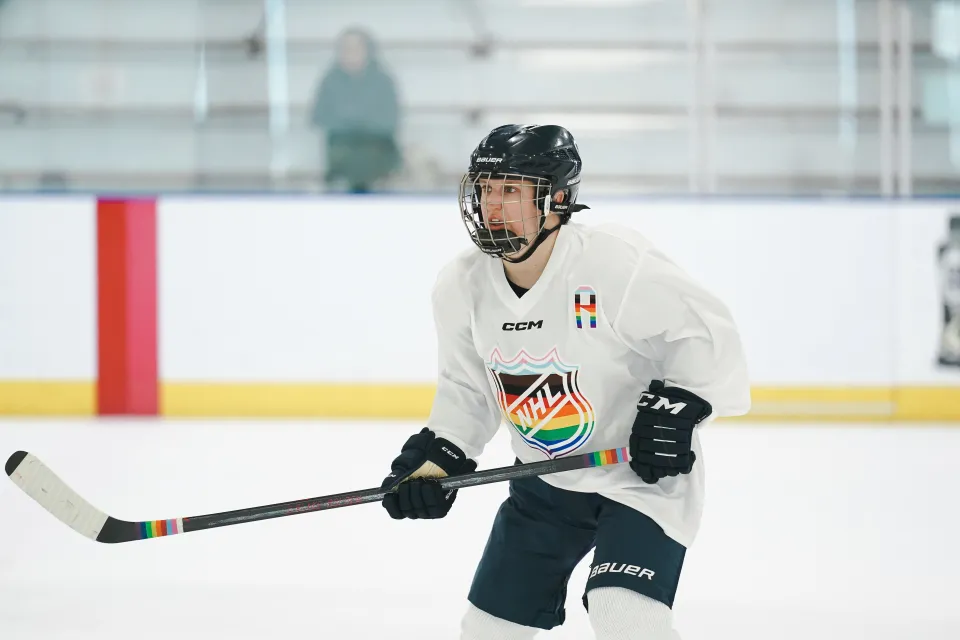 Alex Cothren ice skating in full hockey gear, with a pride-flag jersey.
