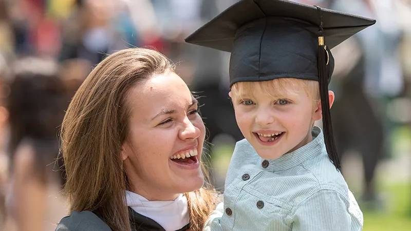Graduating student in their regalia holds a young child wearing their graduation cap