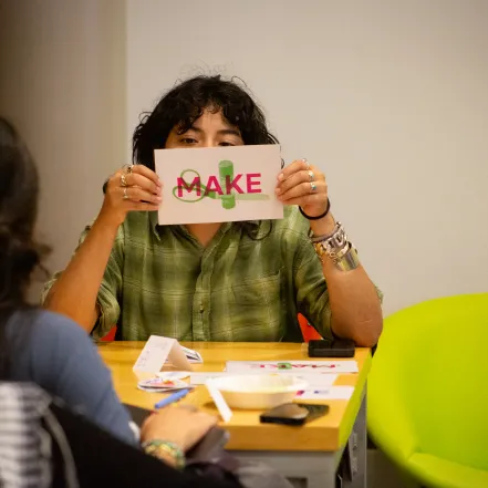 A student holds a paper with the words Make in front of their face while looking at the camera at ZineFest 2024